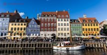Onde ficar em Copenhague: O famoso porto de Copenhague, repleto de casinhas coloridas na beira do mar, com barcos ancorados. O céu está azul. Várias pessoas almoçam na beira do mar, na frente das casinhas coloridas.