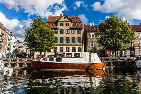 Onde ficar em Copenhague: O bairro de Christianshavn visto do tour do barco pelos canais da cidade. Uma casa amarela de telhas terracota está ao fundo, com um barquinho em frente a ela, flutuando no canal. Árvores estão na beira do canal e o céu está azul com nuvens.