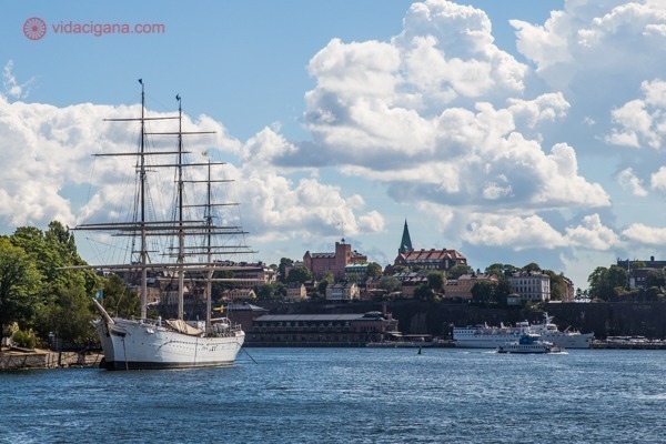 Onde ficar em Estocolmo: O bairro de Sodermalm no fundo, com prédios em colinas, na beira do mar. Um navio antigo está ancorada em frente. O céu está azul com nuvens brancas