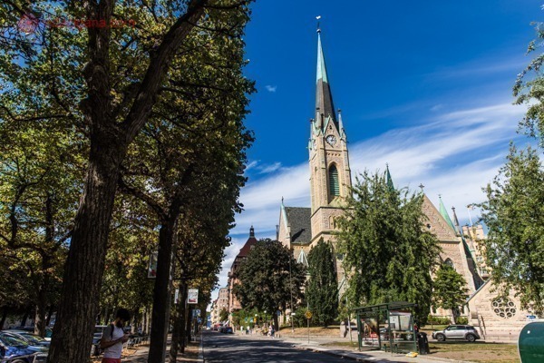 Onde ficar em Estocolmo: o bairro de Östermalm, com uma igreja gótica com torre alta e pontiaguda amarela, árvores verdes no entorno e céu azul com algumas nuvens