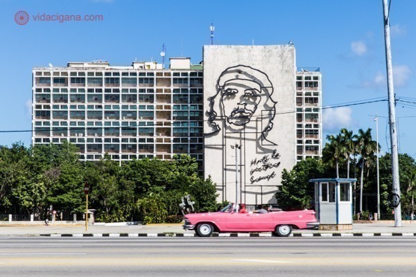Principais pontos turísticos de Cuba: A Praça da Revolução, com o prédio com o rosto de Che Guevara, e um carro antigo cor de rosa passando em frente.