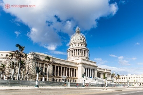 Principais pontos turísticos de Cuba: O Capitólio de Cuba, muito parecido com o de Washington, nos Estados Unidos. Uma prédio branco com uma ala em restauração. O céu está bem azul, com algumas nuvens brancas.