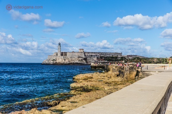Principais pontos turísticos de Cuba: O famoso Malecón, avenida a beira mar de Havana, com a fortaleza ao fundo. O céu está azul, com poucas nuvens brancas. O mar é azul também.