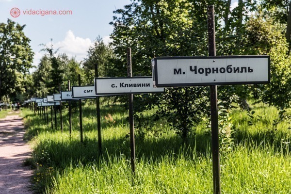 Como visitar Chernobyl: O Beco da Memória e da Esperança, com as placas com os nomes das cidades e vilarejos evacuados durante o desastre. As placas estão fincadas em meio à grama, cercada por árvores.