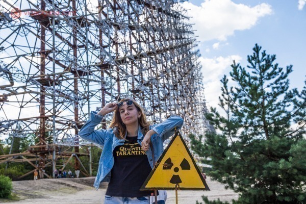 Como visitar Chernobyl: As Torres de Espionagem Duga, altíssimas, no fundo. Uma mulher encostada numa placa indicando radioatividade em primeiro plano.