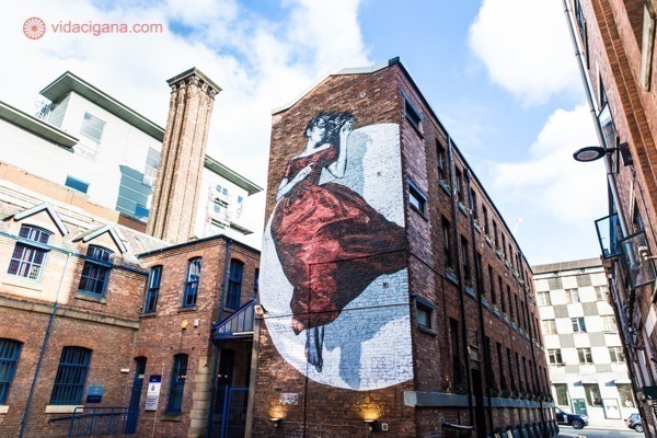 Painel de street art vertical representando uma mulher em vestido longo vermelho. A obra homenageia o movimento das sufragistas que iniciou em Manchester a luta pelo direito ao voto feminino