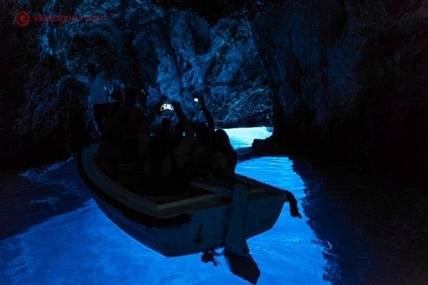 Passeio de barco em Hvar: o interior da caverna azul, com suas águas azuis transparentes e fluorescentes. Um barco passa pela caverna com várias pessoas filmando com seus celulares.