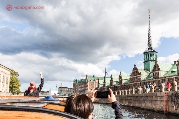 Passeio de barco em Copenhague: foto tirada de dentro do barco, com o guia na frente e um turista com a cabeça do lado de fora do barco tirando uma foto com um celular. Ao fundo, vários prédios famosos a direita
