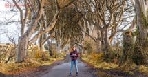 Roteiro Irlanda: A rua na Irlanda do Norte chamada The Dark Hedges, onde filmaram Game of Thrones. A rua é cheia de árvores que criam um arco. Uma mulher está parada no meio da estrada