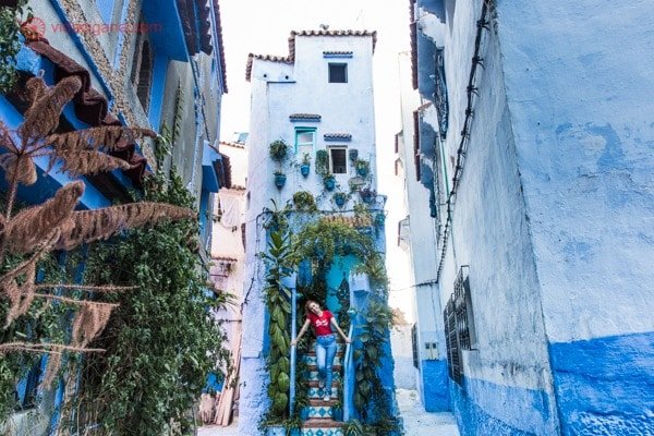 Chefchaouen, a cidade azul do Marrocos: Uma casinha azul bem estreita, como uma torre, no meio de uma ruela bem estreita, tudo azul. Uma mulher com camisa vermelha se encontra nos degraus da casinha