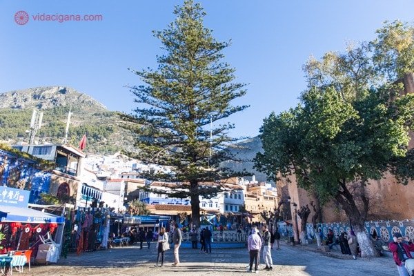 Chefchaouen, a cidade azul do Marrocos: A praça Uta el-Hamman com seu enorme pinheiro no centro, várias pessoas caminhando a sua volta, com vários restaurantes e as montanhas Rife ao fundo