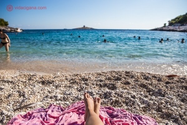 A praia de Podonjo Dol, em Hvar, com sua água cristalina, uma ilha ao fundo, os pés de uma mulher numa canga rosa olhando o mar