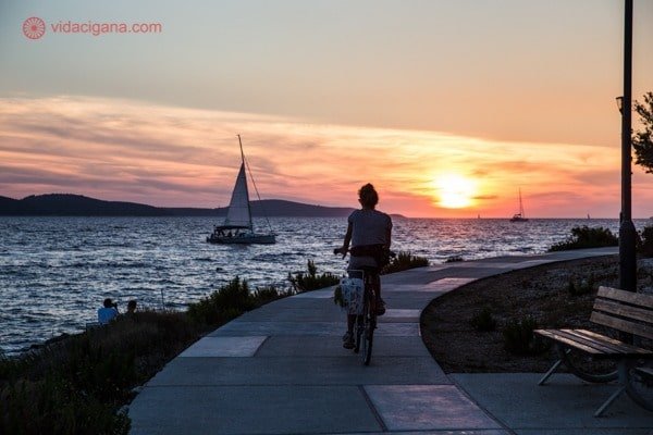 Onde ficar em Hvar: A Hula Hula Beach, com uma mulher na bicicleta andando na calçada enquanto o sol se põe lá no fundo. Um veleiro passa no mar.