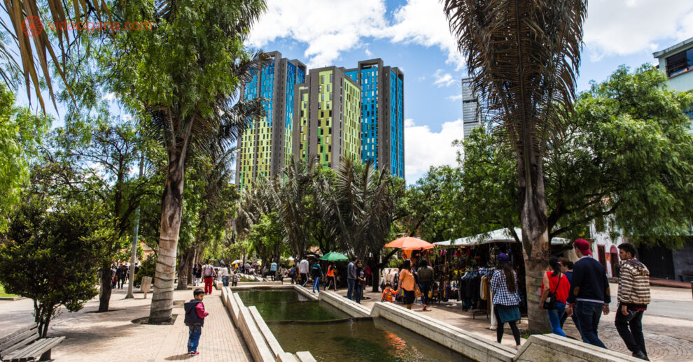 Um parque cheio de árvores em Bogotá com prédios coloridos ao fundo e uma fonte no meio