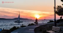 O que fazer em Hvar: uma mulher pedalando em uma bicicleta na frente do mar, na frente do sol se pondo, com um veleiro passando do lado esquerdo da foto. O céu está laranja