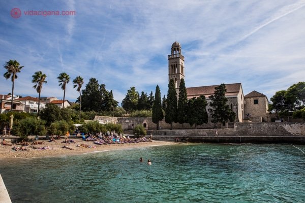 O que fazer em Hvar: O monastério franciscano visto na direita da foto com sua torre do relógio, cercado de árvores e a praia ao lado, com pessoas na areia e nadando