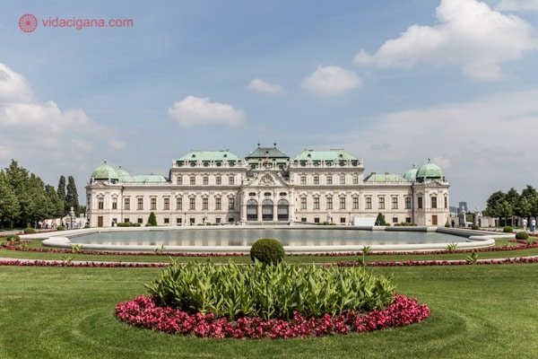 seguro viagem europa: Viena com seu Palácio Belvedere, lindíssimo, com paredes bege e telhado verde, uma enorme fonte e um jardim com flores vermelhas em frente a ele