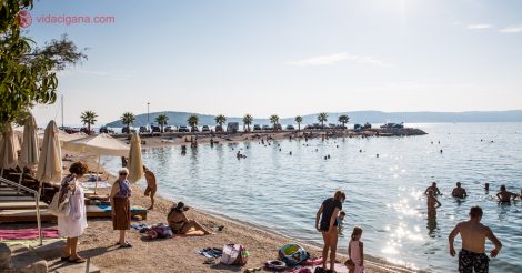 O que fazer em Split: uma praia na Croácia com várias pessoas na areia, adultos, crianças e idosos, com o mar bem calmo e com uma passarela de pedras ao fundo com palmeiras. O céu está azul e o sol brilha a contraluz