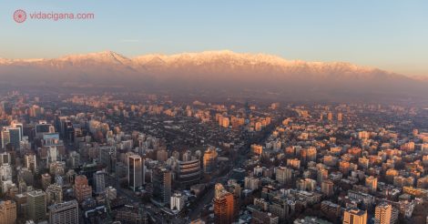 O que fazer em Santiago: Santiago vista do alto do mirante do Sky Costanera, com os prédios embaixo e a Cordilheira dos Andes cheia de neve ao fundo. A foto foi tirada durante o pôr do sol