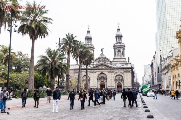 O que fazer em Santiago: A Plaza de Armas no centro de Santiago, com a igreja ao fundo e palmeiras no meio da praça, que está com várias pessoas caminhando