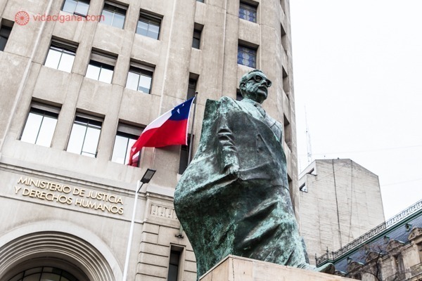 a estátua de Salvador Allende em frente a uma bandeira chilena e ao lado do Palacio de la Moneda