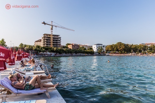 Onde ficar em Split: a praia mais conhecida da cidade, com várias pessoas tomando sol na calçada no entorno do mar azul, com alguns prédios em construção ao fundo