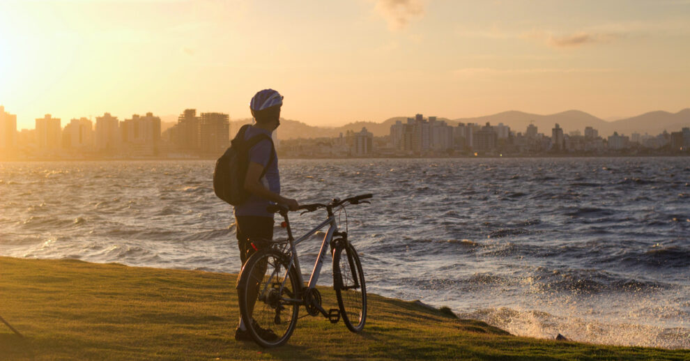 Onde ficar em Florianópolis: Um homem parado na beira do mar com uma bicicleta olhando a cidade ao fundo