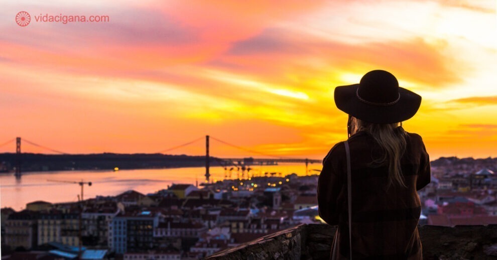 Os 9 Melhores Miradouros de Lisboa, este é o primeiro lugar, o Castelo de São Jorge, com uma mulher usando chapéu de costas vendo o rio Tejo durante o pôr do sol