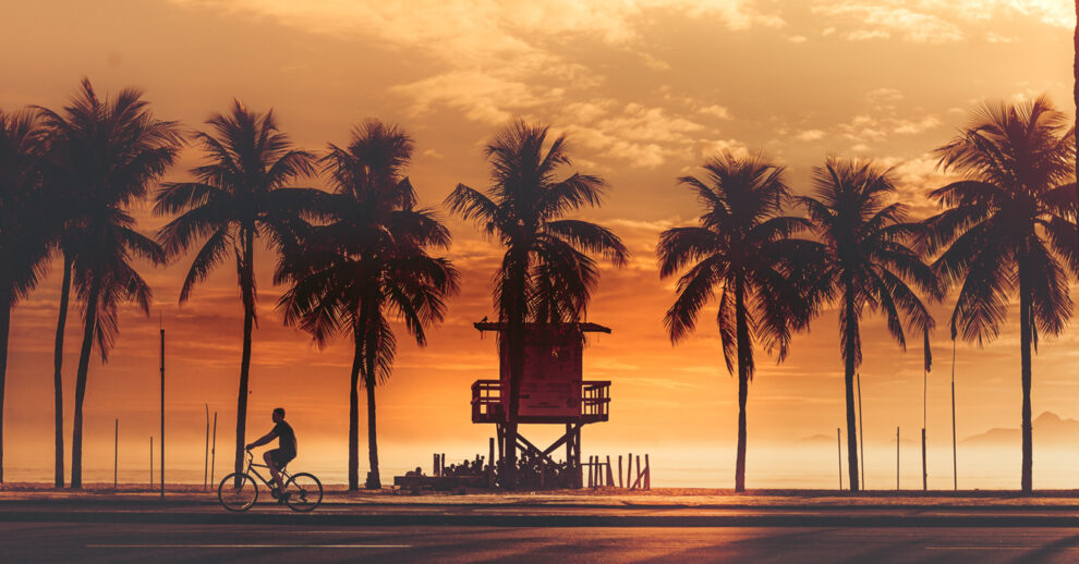 O que fazer em Copacabana: um homem andando de bicicleta no calçadão da praia, perto de um posto e cheio de coqueiros durante o pôr do sol
