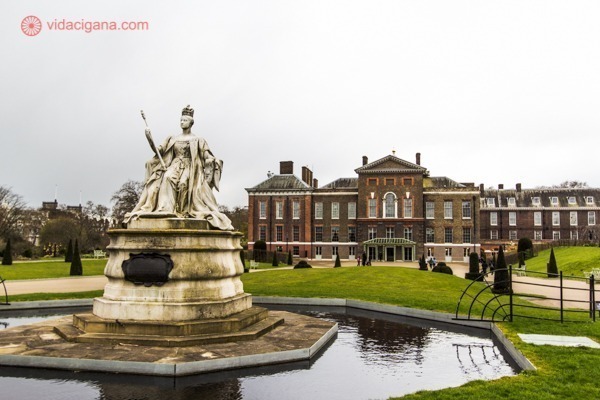 O Palácio de Kensington visto dos Kensington Gardens, com a estátua da rainha Victoria na fonte