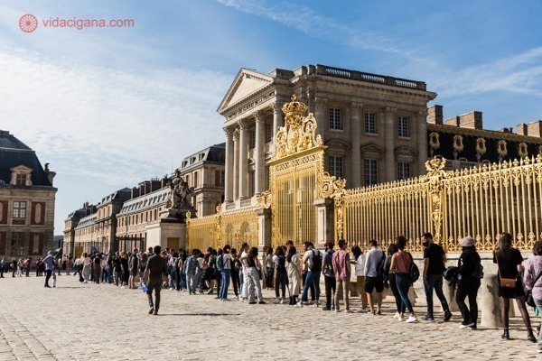 Os portões dourados do Palácio de Versailles com uma fila de pessoas em sua entrada