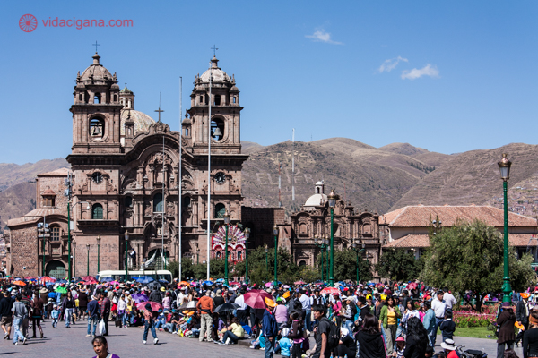 A Catedral de Cusco cheia de pessoas a sua frente, com montanhas atrás, num dia ensolarado