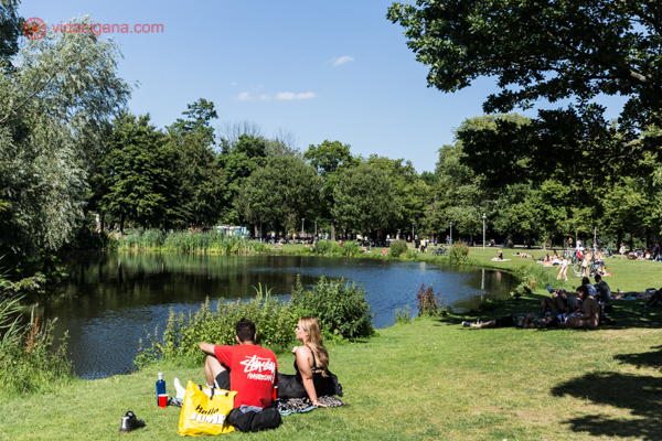 O Vondelpark em um dia de sol, cheio de verde e muitas pessoas sentadas na beira do lago