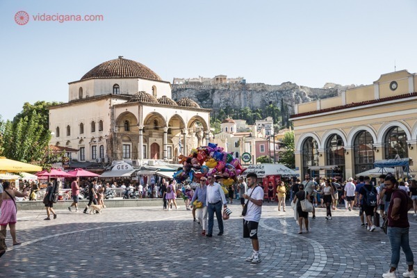 A Praça Monastiriki com vendedores de balões e muitos turistas, com a Acrópole ao fundo e ao alto