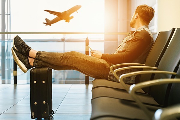 Coberturas de seguro viagem: Um homem sentado em um banco de aeroporto, com os pés apoiados na mala e observando um avião voar lá fora