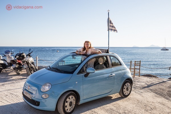 Uma mulher num carro conversível azul claro em frente ao mar com a bandeira da Grécia atrás