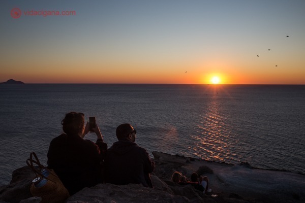 Duas pessoas vendo o pôr do sol no mar e tirando fotos com o celular