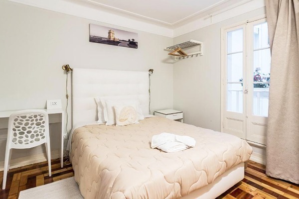 Uma cama de casal clara com cobertor bege, num quarto com escrivaninha e portas que dão para uma varanda