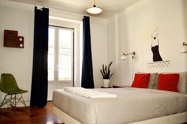Uma cama cinza com travesseiros vermelhos, portas que dão para uma sacada e cortinhas pretas