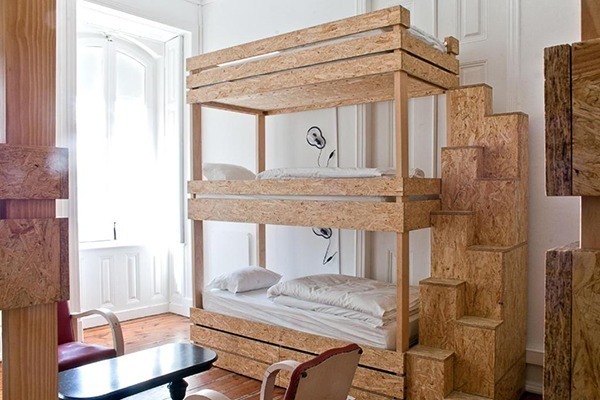 Uma beliche de hostel, feita de madeira reciclata, com lençóis claros e perto da janela