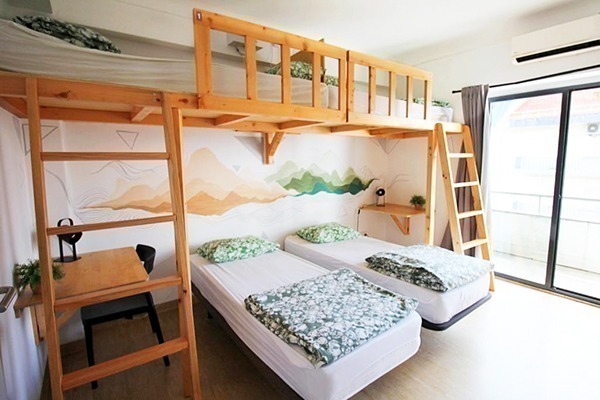 Duas camas de solteito e 2 beleches só na parte de cima, com lençóis brancos e verdes, pintura na cabeceira das camas de montanhas verdes e laranjas. Portas que dão pra sacada