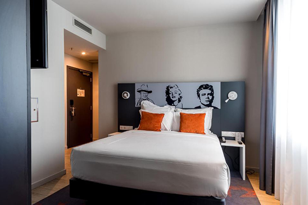 Um quarto com paredes brancas, cama com bom tamanho, cabeceira com estrelas de cinema como Marilyn Monroe e James Dean