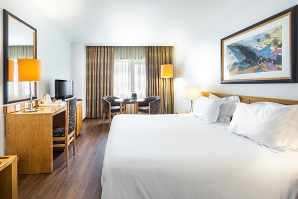 Um quarto de hotel bem iluminado com janelas grandes, cama enorme branca, com televisão, uma mesinha com 2 poltronas