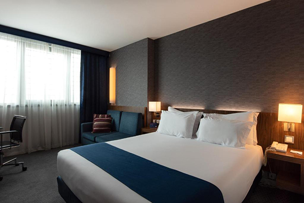 Um quarto de hotel escuro, com paredes negras, lençóis da cama brancos e sofá azul escuro