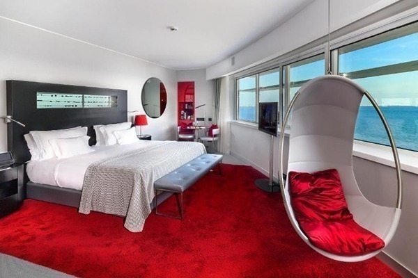 Um quarto com visual moderno, com cadeira suspensa do teto, com almofadas vermelhas, cama com cabeceira preta e janela com vista para o rio