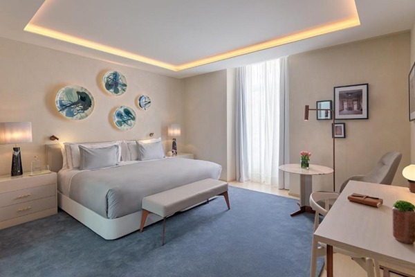 Um quarto de hotel com uma cama bem grande com pratos decorativos na cabeceira, janela bem iluminada, 2 mesas com poltrona