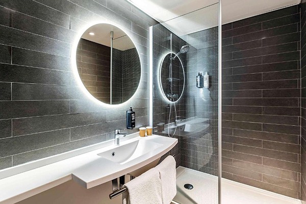 Banheiro com azulejos pretos, espelho redondo com iluminação atrá dele, pia branca