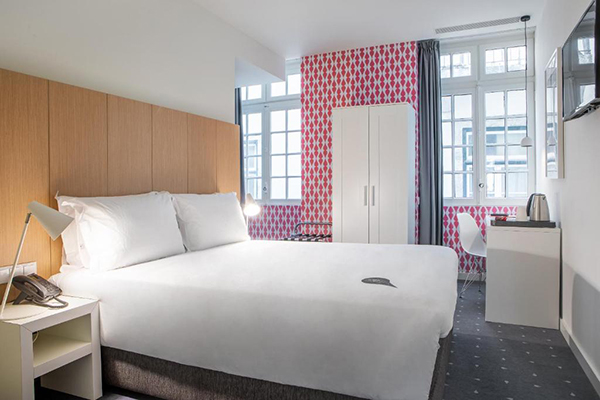 Um quarto pequeno, mas confortável, com uma cama grande branca e papel de parede vermelho e branco