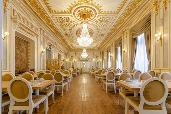 Um restaurante de hotel todo branco e dourado, cheio de cadeiras confortáveis e acolchoadas, e o teto parecendo o Palácio de Versailles