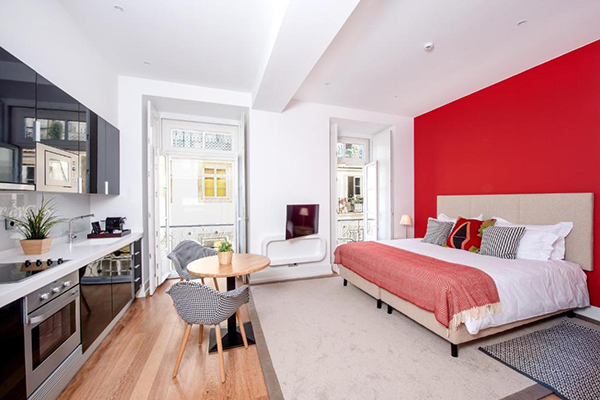 Um quarto branco com a parede da cama vermelha, cozinha preta e uma mesa com 2 poltronas.